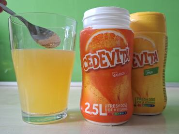 Egy kéz Cedevita üdítőitalport kanalaz egy pohár vízbe.