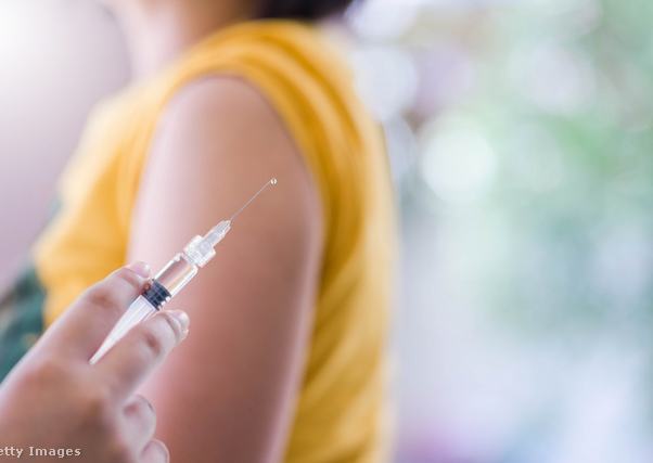 Injekciós tűt tart egy orvos, a háttérben a beteg feltűrt póló ujjal vár