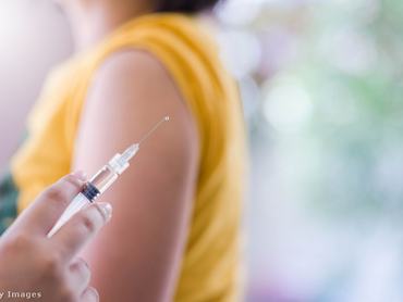 Injekciós tűt tart egy orvos, a háttérben a beteg feltűrt póló ujjal vár