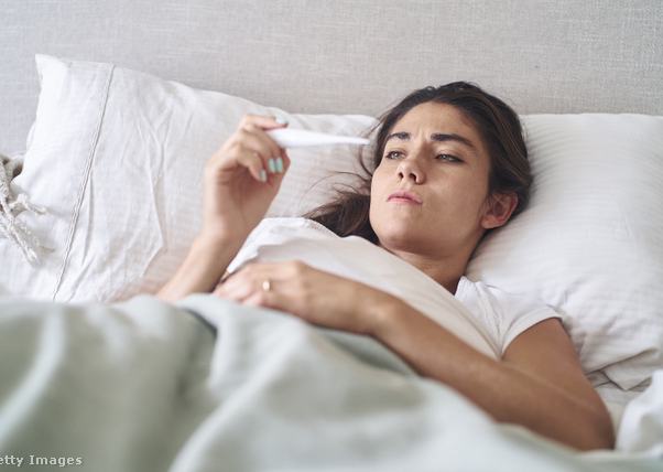 egy beteg nő fekszik a kórházi ágyon, a lázmérőt nézi