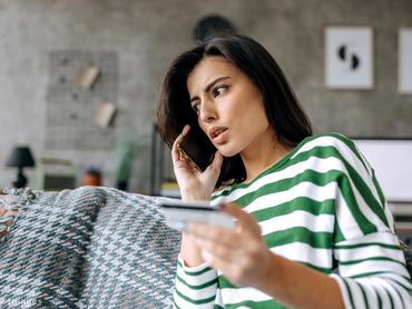 kétségbeesett nő telefonál, másik kezében bankkártyát tart, miközben a kanapén ül