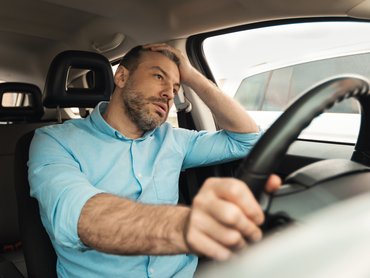 Ideges férfi ül az autó első ülésén, a fejét fogja vezetés közben, másik keze a kormányon.