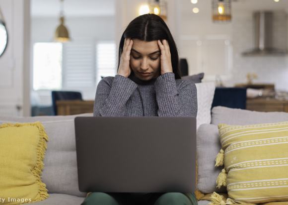Fiatal nő a fejét fogva ül a laptop előtt a kanapén
