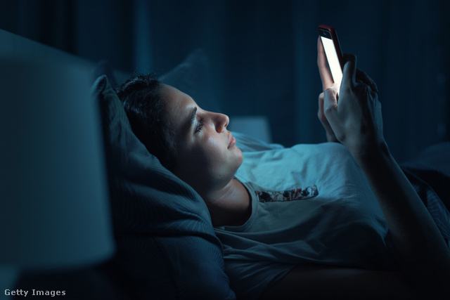 Egy nő sötétben, az ágyban fekve nézi a világító telefonját