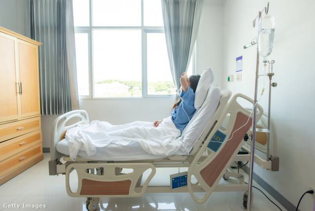 egy hosszú barna hajú beteg kék kórházi ruhában fekszik a kórházi ágyban, kezeit magadba emeli, mellette infúzió, a nap süt a szobába