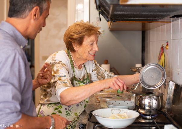 egy idős hölgy és egy fiatalabb férfi együtt főza konyhában, a nő a tűzhely mellett áll, a fedőt emeli