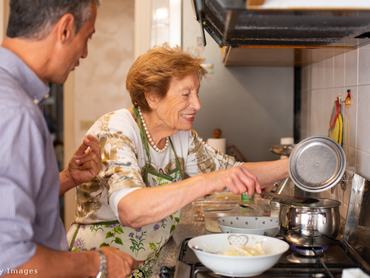 egy idős hölgy és egy fiatalabb férfi együtt főza konyhában, a nő a tűzhely mellett áll, a fedőt emeli