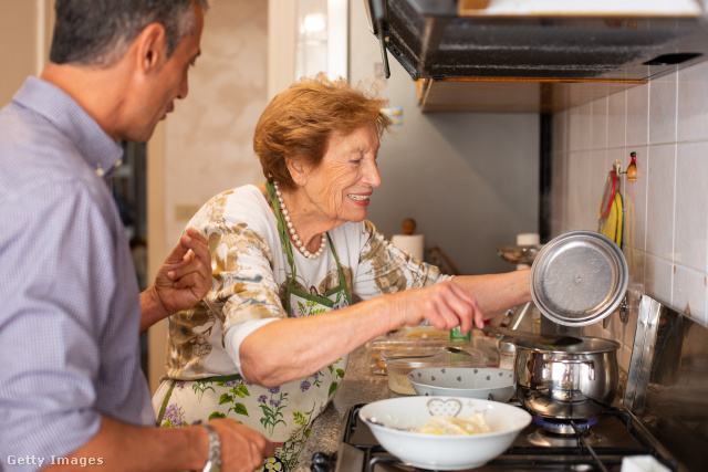 egy idős hölgy és egy fiatalabb férfi együtt főza konyhában, a nő a tűzhely mellett áll, a fedőt emeli