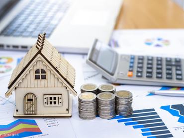 Fából készült házmakett, pénz, toll, számológép és statisztikai adatok hevernek az asztalon