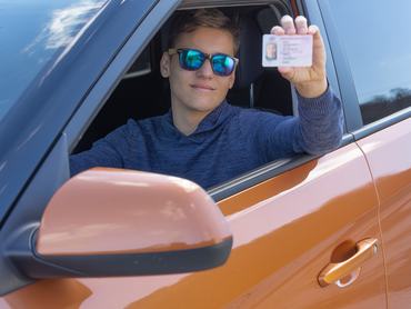 Happy teenager is showing his driving license from open car window. Driving school concept