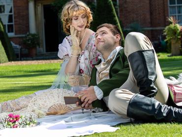 Young lovers dressed in vintage clothing sitting on picnic blanket. Gentleman looking up at his partner while holding book