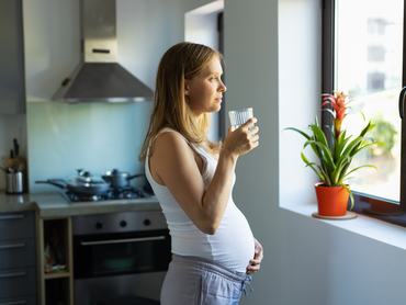 Töprengő kismama áll a konyhában pohár vízzel