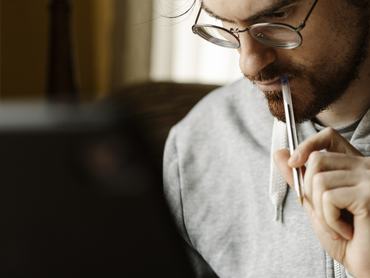 Concentrated thoughtful adult male journalist in casual wear and eyeglasses touching lips with pen while reading article during work at home