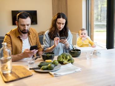 Fiatal szülők okos telefonjukba merülve ülnek ebédidőben az asztalnál egyéves babájukkal.