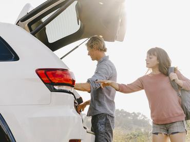 Fiatal pár áll fehér autójuk nyitott csomagtartója előtt, a férfi pakol, a nő valamire mutat az autó belsejében.