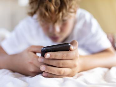 kamasz fiú hasal a vetett ágyon telefonnal a kezében