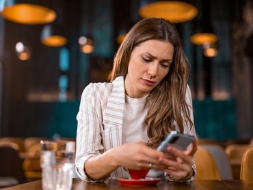 Aggódó és elégedetlen fiatal nő rossz híreket olvas a telefonján, miközben egy kávézóban ül.