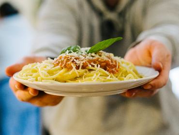 Közeli kép egy friss tál tésztáról szósszal és sajttal, amit valaki felé tart.