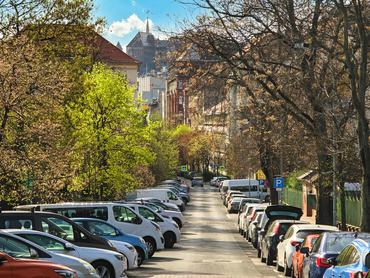 Egy csendes városi utca, melyet mindkét oldalon parkoló autók szegélyeznek, kora tavasszal virágzó fák veszik körül. A látvány a békés környék hangulatát ragadja meg az építészet és a természetes zöld növényzet keverékével. Helyszín: Budapest, Magyarország