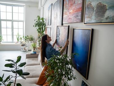 Woman painter hanging newly painted modern framed painting on wall in bright room art workshop with indoor plants. Actual solutions by decoration in contemporary interior design. Artists cosy home