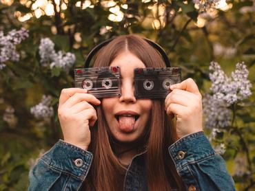 Happy young woman holding audio cassettes in front of her eyes while listening music in headphones during spring