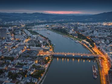 Aerial View of Linz, Austria, at blue hour, showing the illuminated bridges over the Danube River, the city skyline, and surrounding hills under a soft morning sky. Linz in Upper Austria, Europe.