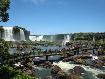 Tourists walk along the panoramic footbridges of Iguaçu National Park, gazing at the magnificent Iguaçu Falls in Foz do Iguaçu, Brazil. The photo captures the immense power and natural beauty of one of the largest waterfall systems in the world, surrounded by lush subtropical forest under a bright blue sky.