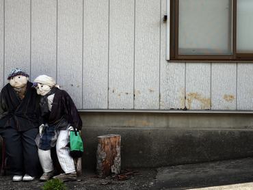 két madárijesztő-figura kuporog egymás mellett egy japán faluban