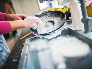 Closeup of unrecognizable woman doing dishes with lots of dirty plates and pots waiting aside. She's cleaning a black non stick cooking pan.