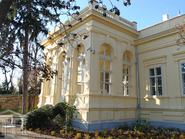 A A balatonfüredi Jókai Mór Emlékház kívülről. Az előtérben egy fa lombja, a ház világos színűre van festve.