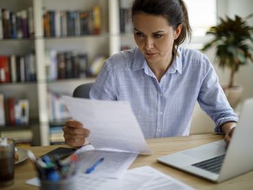 Egy nő az adózási papírjait nézi miközben otthon dolgozik a számítógépen