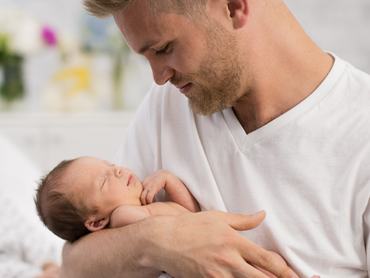 Egy szerető apa ringatja újszülöttjét, miközben csendes, meghitt pillanatokat élnek át a gondoskodás és a kötődés jegyében. A meleg fény és a nyugodt környezet biztonságot, kényelmet és a család örömét sugározza.