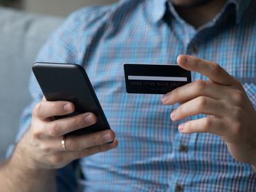 Egy férfi a mobiltelefonját és a bankkártyáját tartja a kezében online vásárlás közben