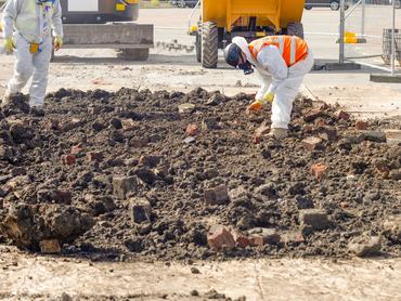 Két védőruházatban dolgozó munkás eltávolítja az azbesztet a téglákkal teli, szennyezett talajból egy építkezésen