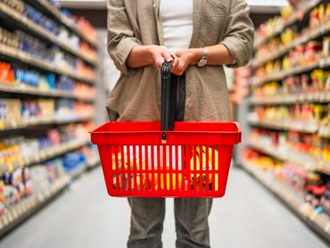Egy nő élelmiszert vásárol a szupermarketben: egy étellel teli piros kosarat tart a kezében, és a folyosón sétál