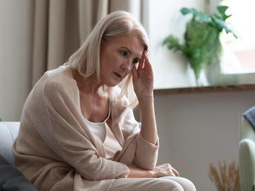 Frusztrált, középkorú nő ül otthon a kanapén, és a fejét fogja. Stresszes, zaklatott, érett nő, aki személyes gondjain vagy egészségügyi problémáin töpreng. Mentális zavar, memóriavesztés, demencia.