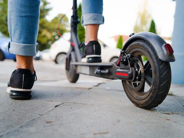 Közelkép egy nő lábáról és lábfejéről, aki nyári napon, hátulról nézve, farmerban és tornacipőben áll egy elektromos rolleren a járdán