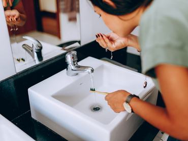 Egy nő a mosdókagyló fölé hajolva vizet vesz a szájába fogmosás után, miközben a kezében fogkefét tart