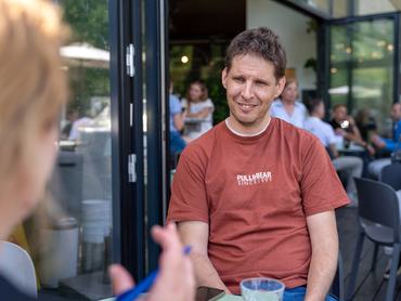 Geri Ádám, a gasztrorégész interjú közben. Téglavörös, rövidujjú pólót visel, a háttérben a kávézóban emberek üldögélnek, az előtérben az újságíró haja.