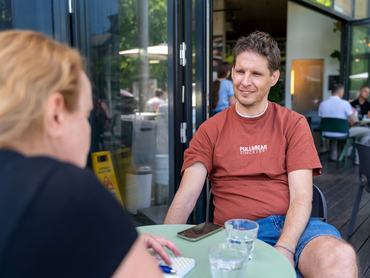 Geri Ádám gasztrorégész, piros pólóban és kék rövidnadrágban. Az előtérben az újságírónő hátulról, a háttérben a kávézóban emberek