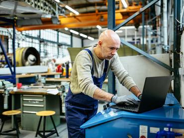 Egy idősebb repülőgép-szerelő egy hatalmas repülőgép-hangárban dolgozik egy laptopon. Munkaruhát visel, például munkaruhát, míg a háttérben gépek és motorok láthatók.