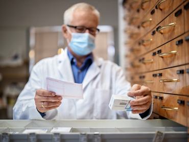 Férfi gyógyszerész maszkban a receptre felírt gyógyszert keresi a patikai fiókokban