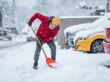 Egy sárga sapkát és piros kabátot viselő férfi lapáttal szorgalmasan takarítja a havat a felhajtóról, miközben a közelben autók állnak