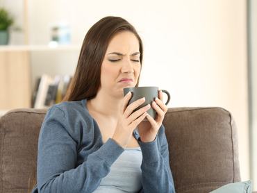 Egy nő a kanapén ülve fintorog mert rossz ízű az itala, amit a kezében tart