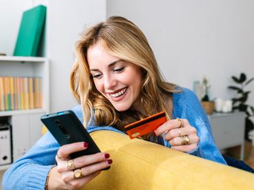 Boldog, fiatal, szőke nő, aki bankkártyát tart a kezében, miközben otthon egy mobilalkalmazáson keresztül online vásárol.
