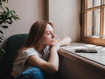 Egy szomorú, vörös hajú tinédzser lány az ablak mellett ül, a telefonját bámulja, és üzenetre vár