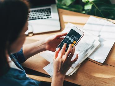 Közelkép egy üzletasszonyról, aki számológépet és laptopot használ pénzügyi számítások elvégzéséhez egy irodai faasztalon, üzleti munkakörnyezetben; adózás, könyvelés, statisztika és elemző kutatás témakörében