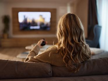 Hátulnézet egy nőről, aki a nappali kanapéján választja ki, mit nézzen a tévében.