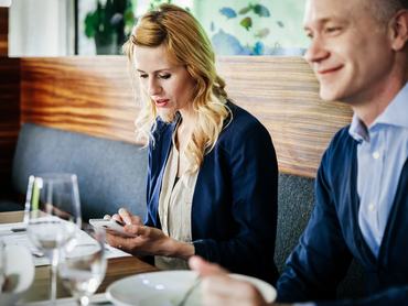 Egy elegánsan öltözött nő, aki üzleti partnereivel együtt ebédel egy étteremben, miközben okostelefonját használja.