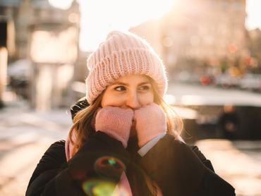 Egy fiatal nő sapkában és kesztyűben, a téli hidegben az utcán állva melegíti a kezét a városban